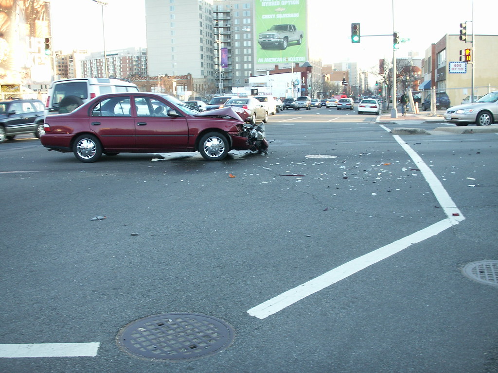 Maroon sedan with crushed front end stopped in a city intersection, debris scattered across the crosswalk
